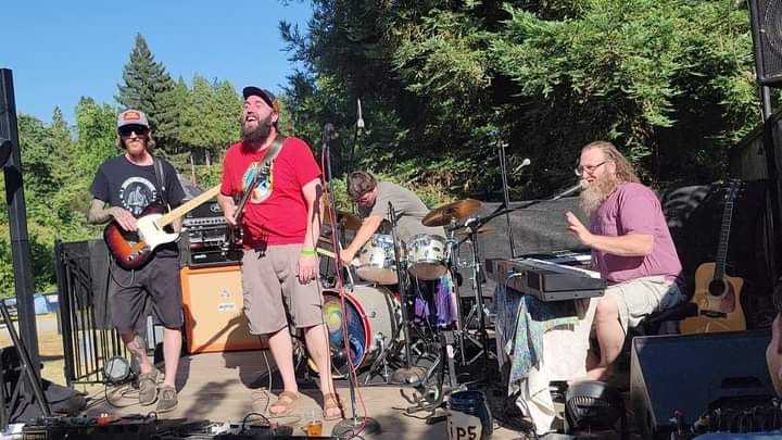 Band performing outdoors with guitars, drums, and keyboard under sunny sky.